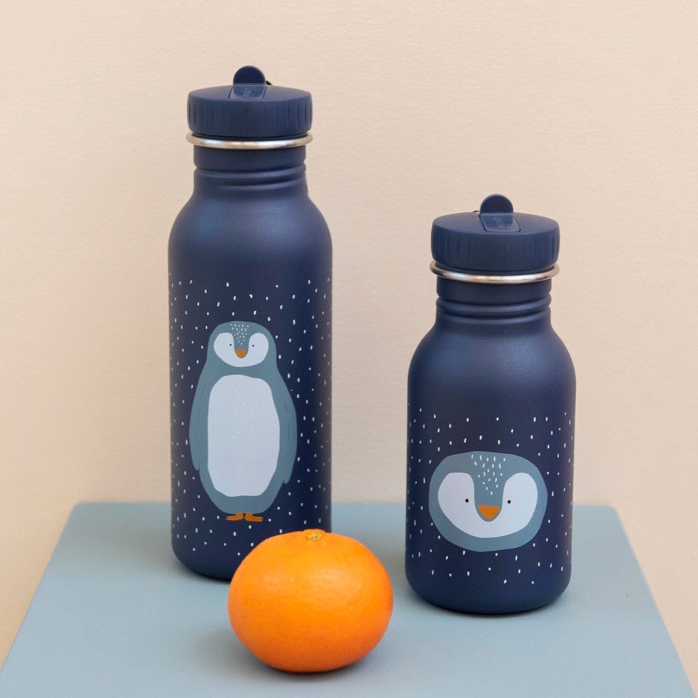 1. Two Trixie Baby Mr Penguin stainless steel bottles in navy with penguin design, placed on a table with an orange, showcasing different sizes