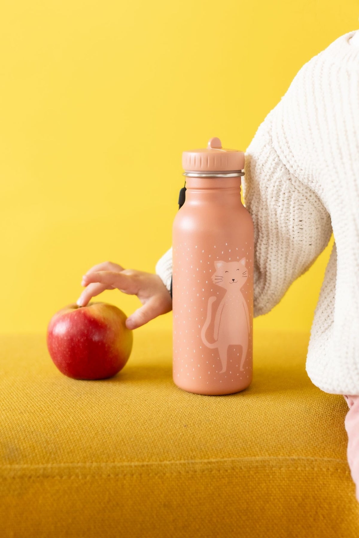 1. Child's hand reaching for apple next to Trixie Baby Mrs Cat stainless steel bottle on yellow surface, against bright yellow background