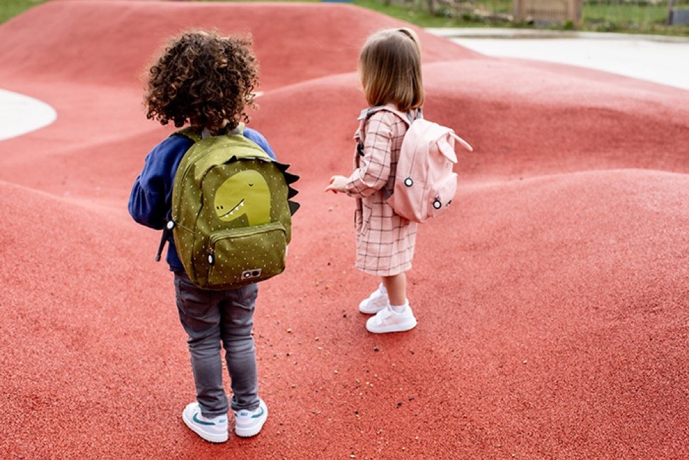 1. Two children wearing backpacks, one with green Mr Dino backpack and the other with pink backpack, standing on red playground surface