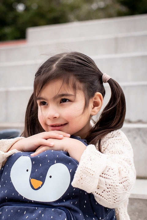 3. Young girl with pigtails holding navy blue Mr Penguin backpack, sitting on outdoor steps, smiling and looking away