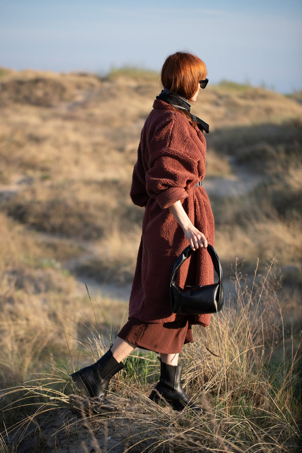 4. Woman with PYKOK Soft Leather Baguette Handbag Luna in black, walking through grassy landscape
