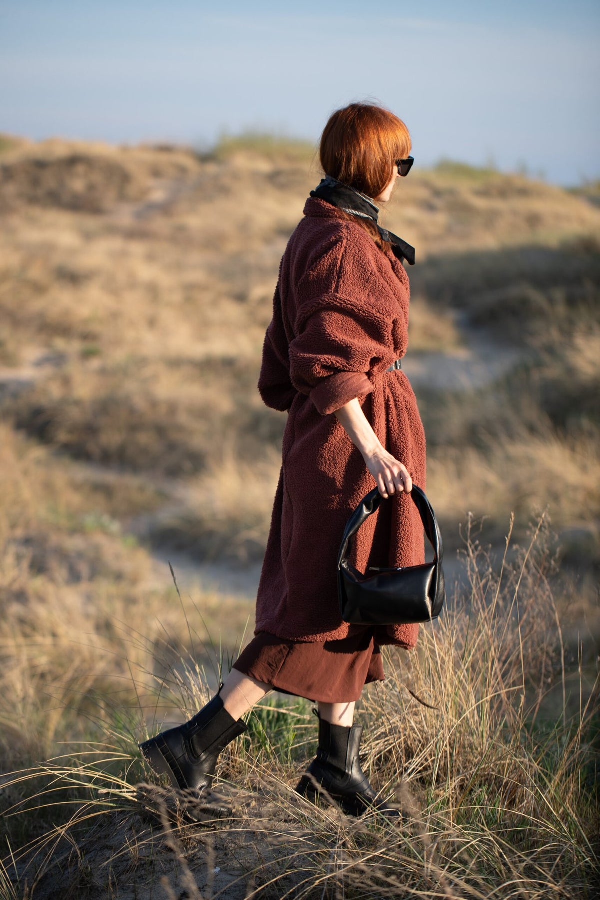 4. Woman with PYKOK Soft Leather Baguette Handbag Luna in black, walking through grassy landscape