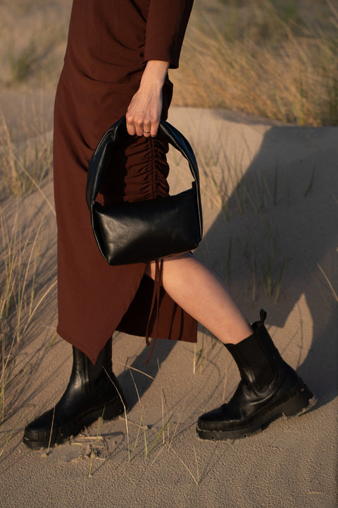 2. Woman carrying PYKOK Soft Leather Baguette Handbag Luna in black, walking on sandy terrain
