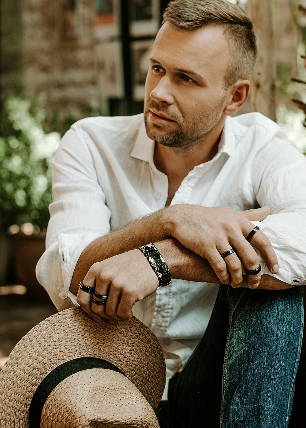 3. Man in white shirt and jeans wearing multiple black ceramic rings and bracelet by Leomani, sitting outdoors with a straw hat