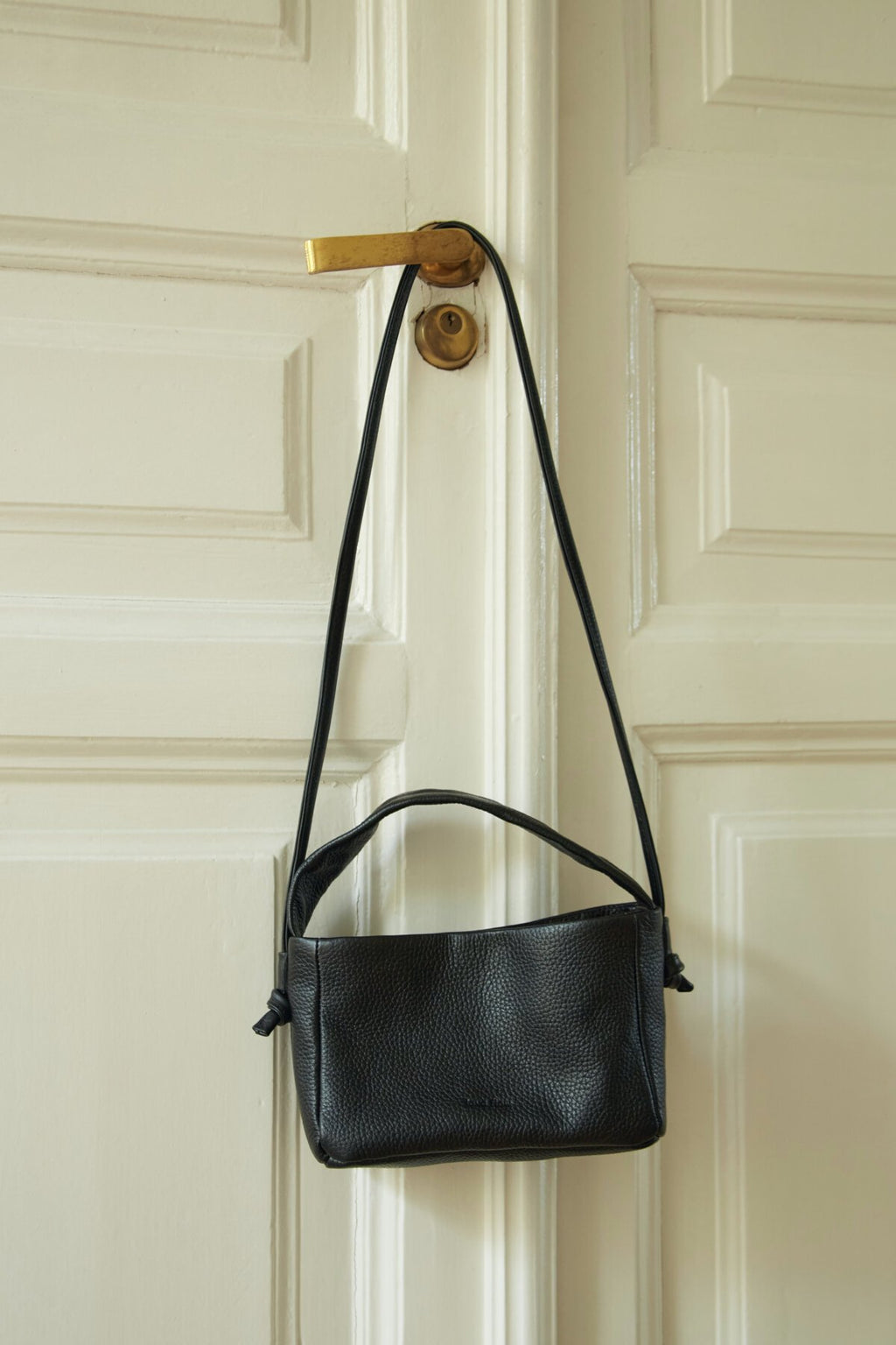 5. Lava Flow Crossbody Bag MERU in black hanging on a door handle, highlighting long strap and Italian leather