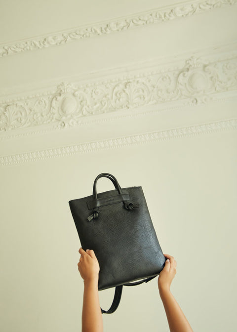 2. Hands holding up Lava Flow ALICE ARM black leather backpack against ornate wall, highlighting knot details