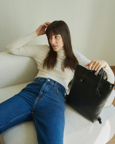 1. Woman sitting on a couch holding Lava Flow ALICE ARM black leather backpack, showcasing minimalist design