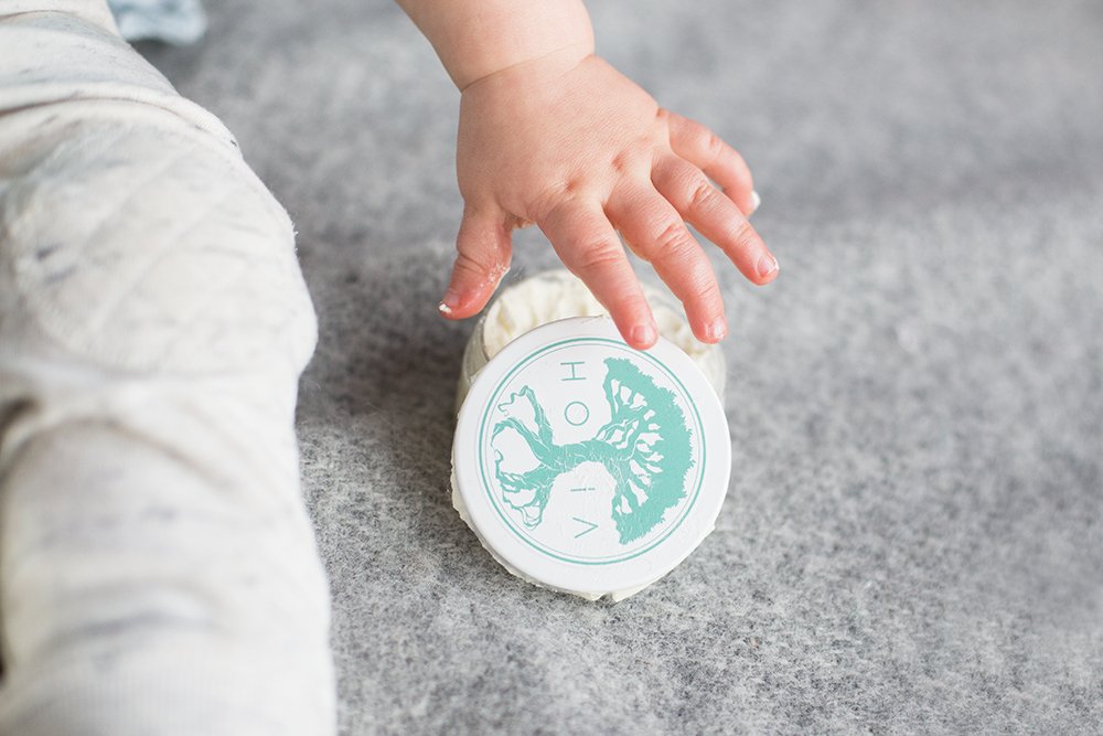 1. Baby's hand reaching for HOIA homespa Baby Cream jar on soft surface, highlighting gentle care for sensitive skin