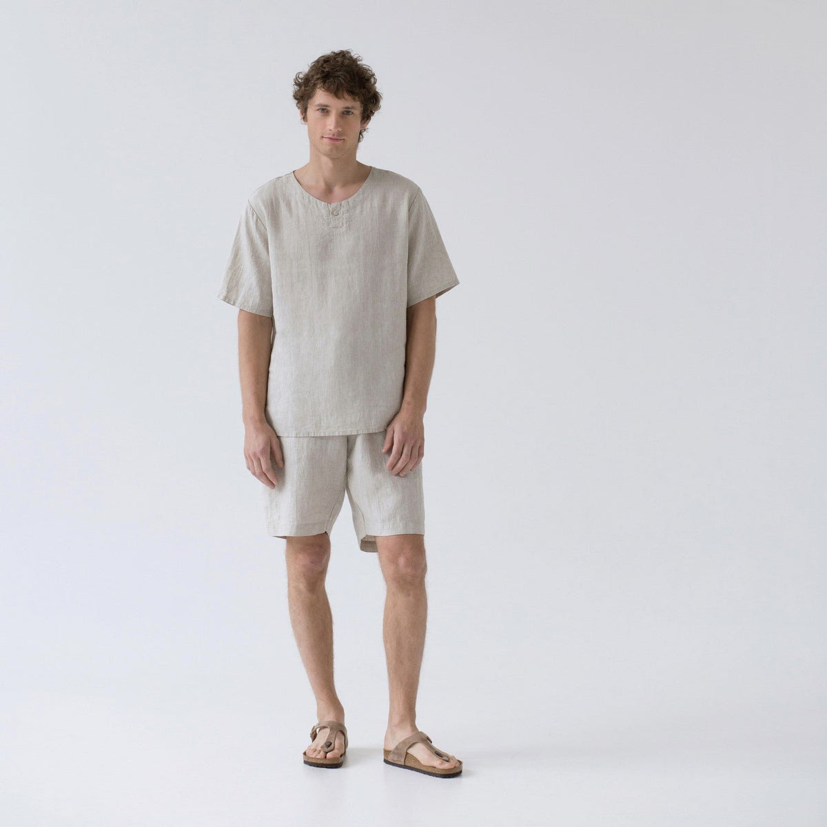 1. Man wearing beige linen pyjama set with short sleeves and shorts, standing in studio setting