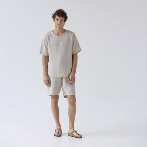 1. Man wearing beige linen pyjama set with short sleeves and shorts, standing in studio setting