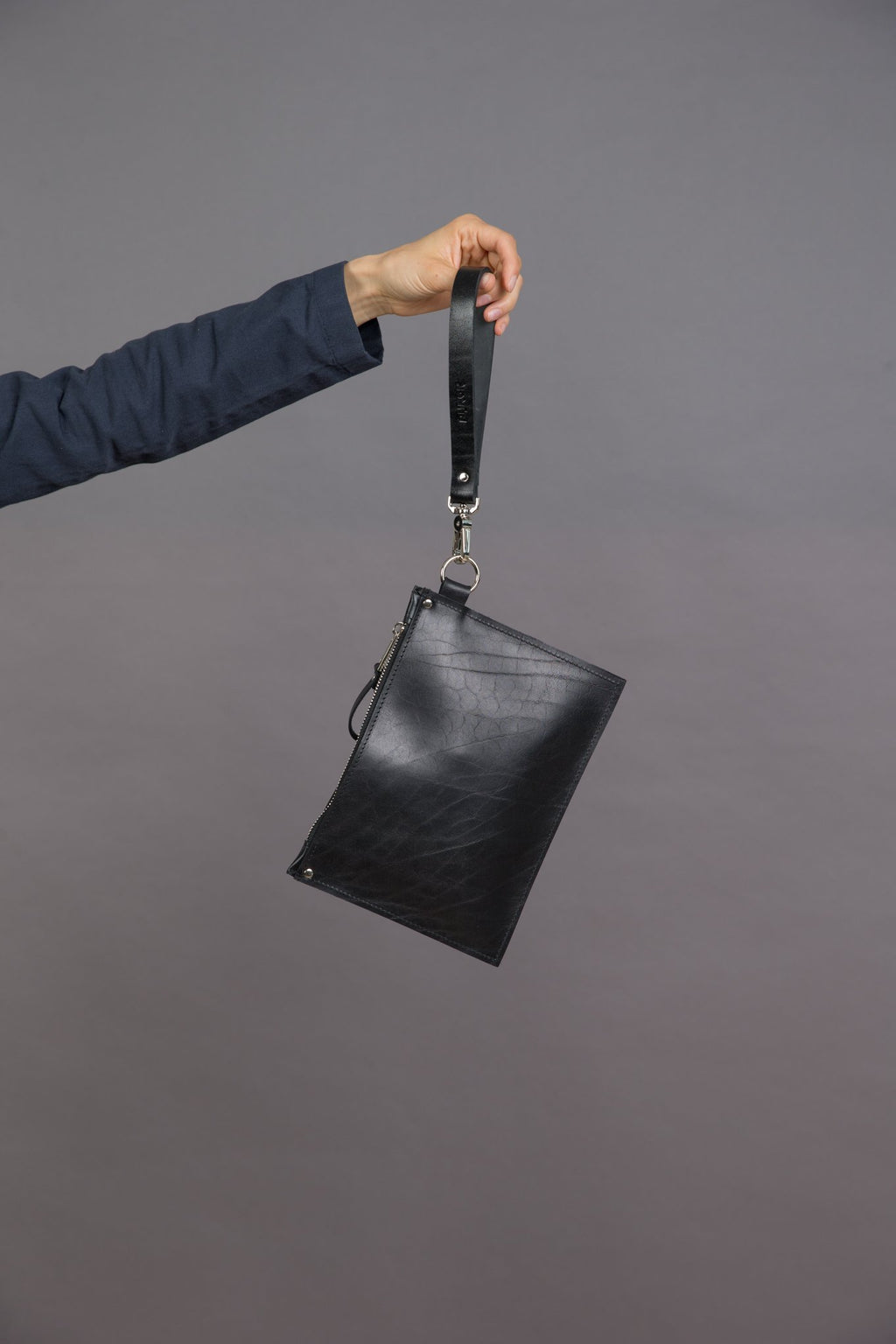 1. Women's black leather Clutch Nina by PYKOK with wrist strap, held in hand against a gray background