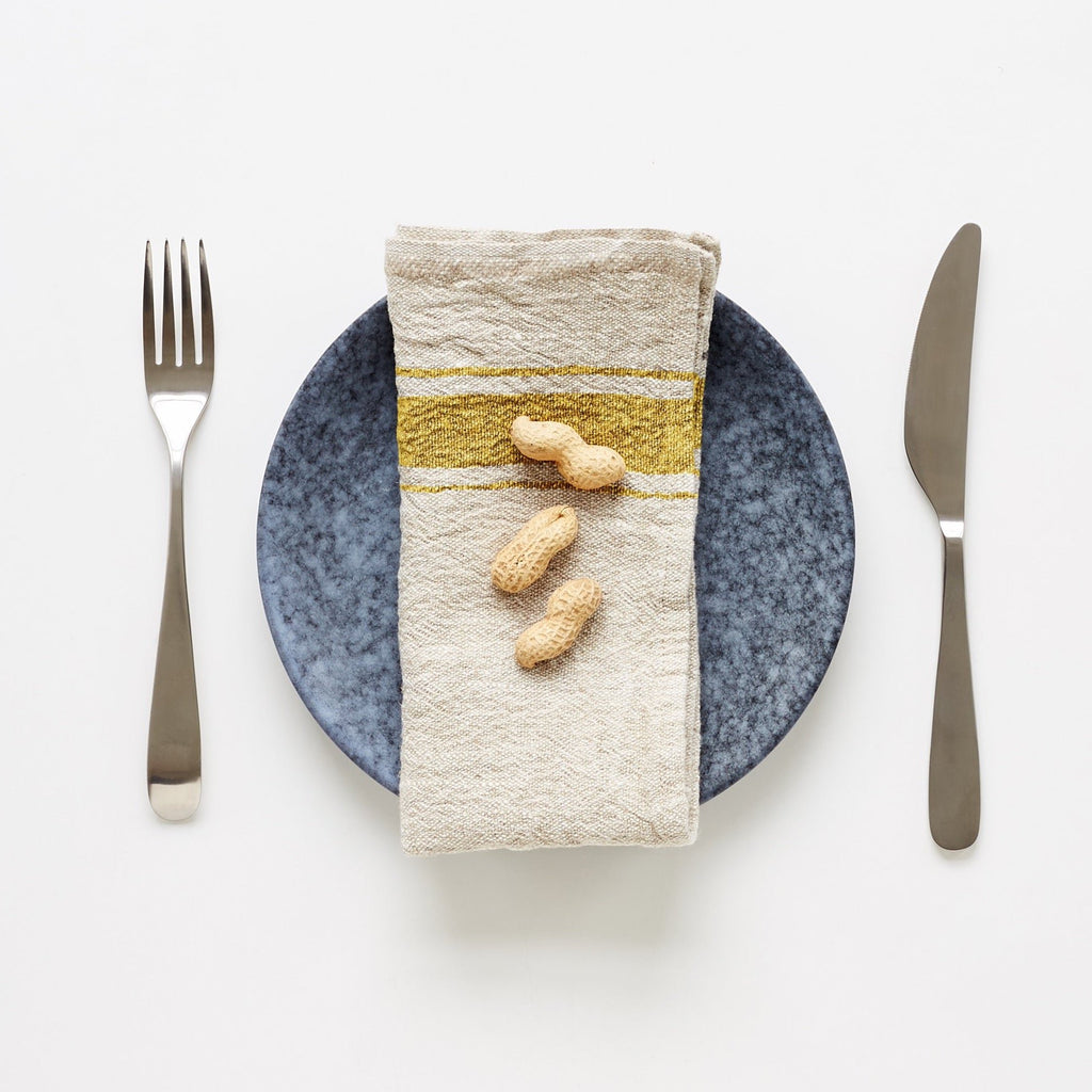 1. Mustard striped linen napkin on blue plate with peanuts, set with fork and knife on white background