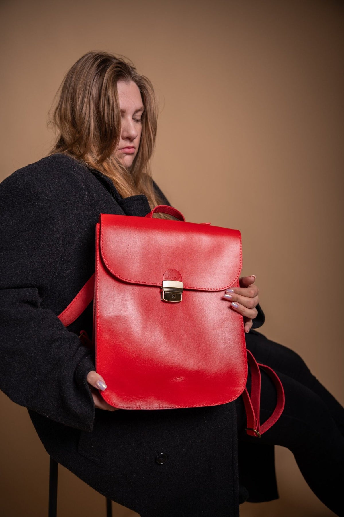2. Woman holding Zelma Kraft red leather saddle backpack, shown in a studio setting with a black coat