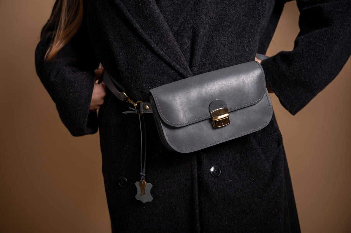 1. Women's grey natural leather saddle bag mini by Zelma Kraft worn with black coat, showcasing adjustable strap and buckle closure