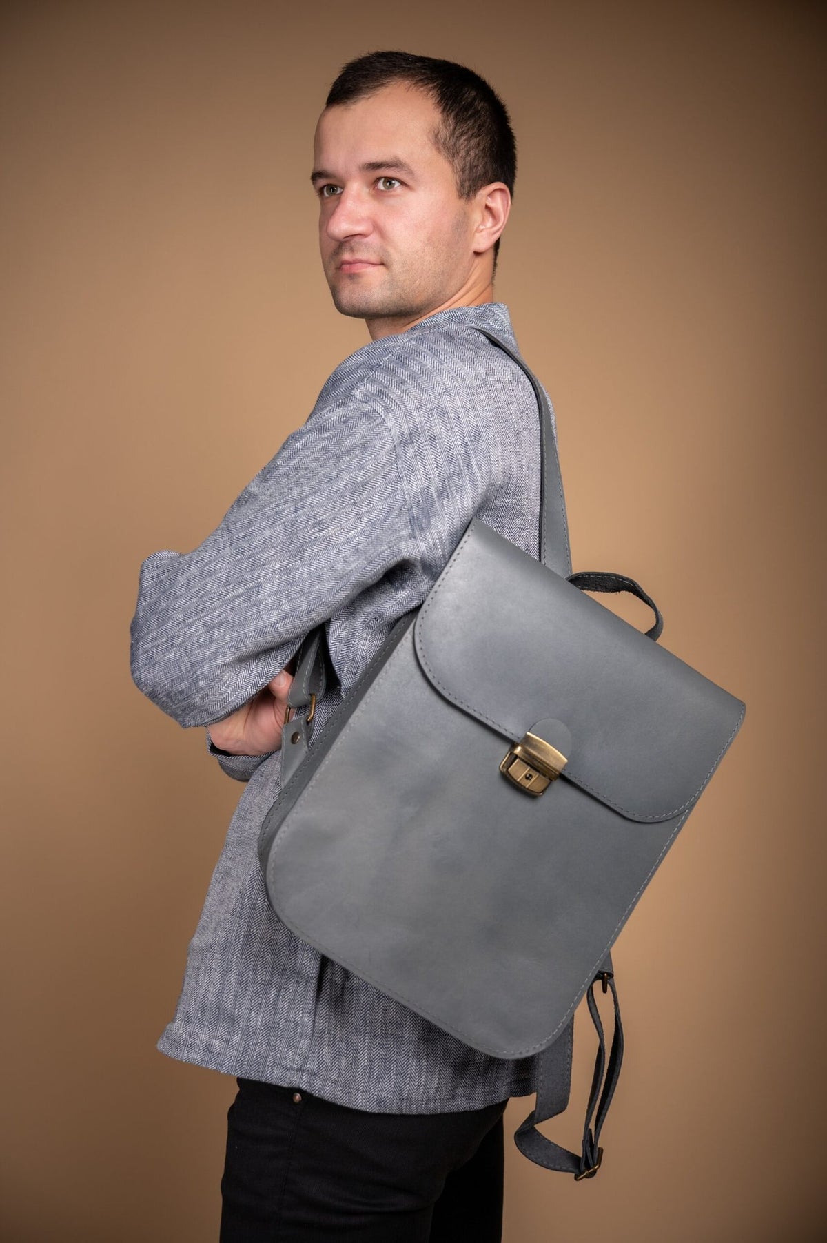 3. Men's grey Zelma Kraft leather saddle backpack worn over shoulder, shown in studio setting