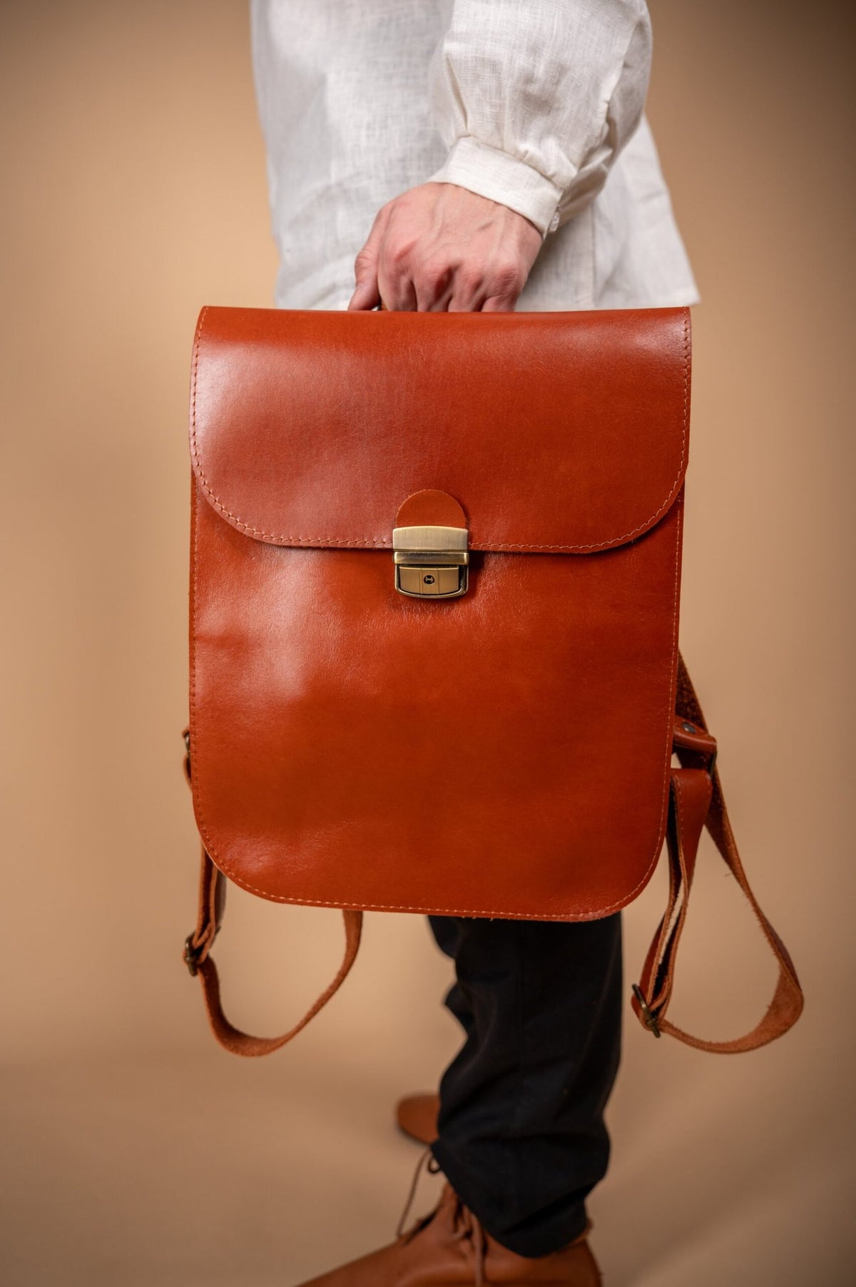 5. Close-up of Zelma Kraft light brown leather backpack held by man, highlighting front design and clasp in studio setting