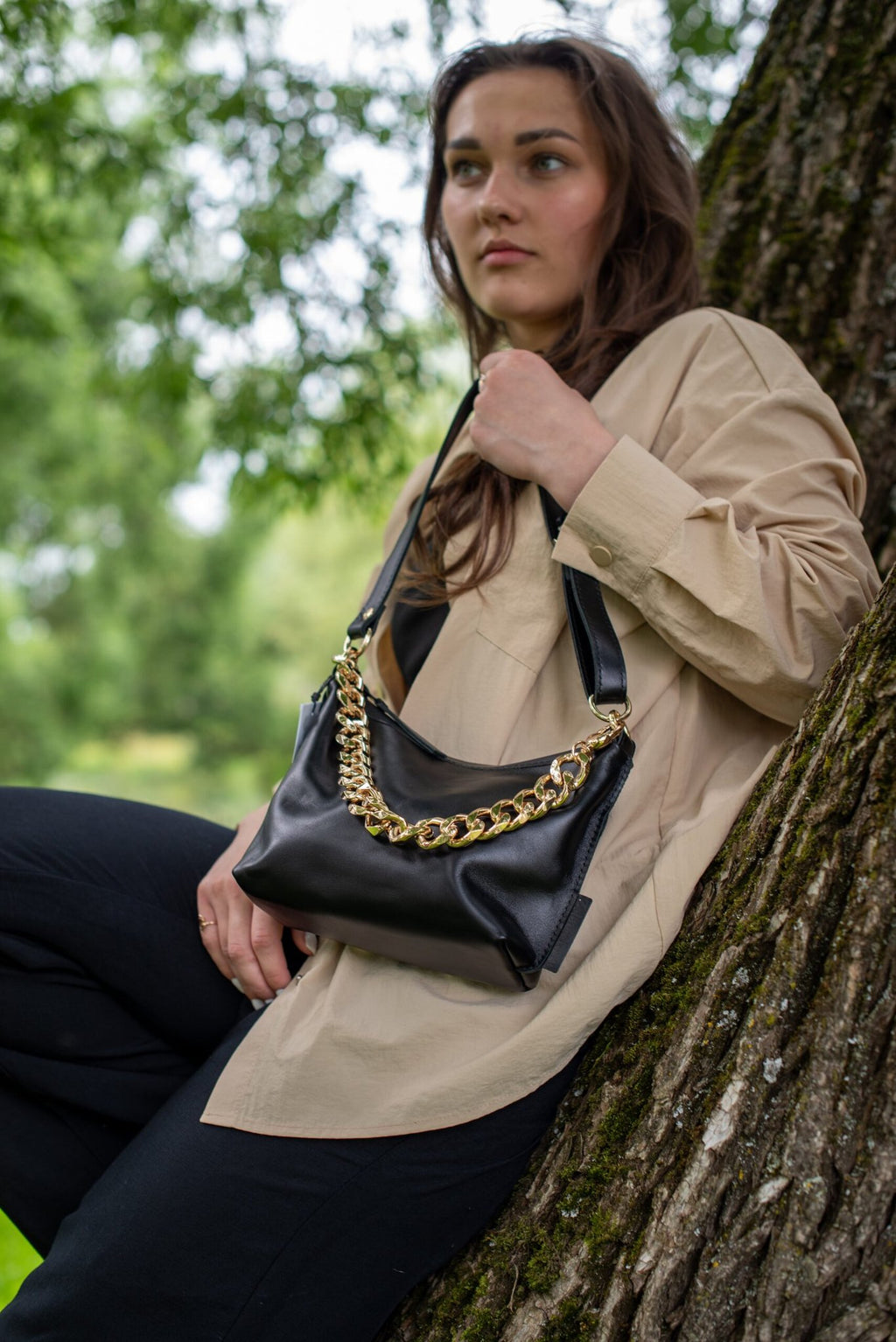 3. Woman sitting by tree with Zelma Kraft black leather hobo bag featuring gold chain