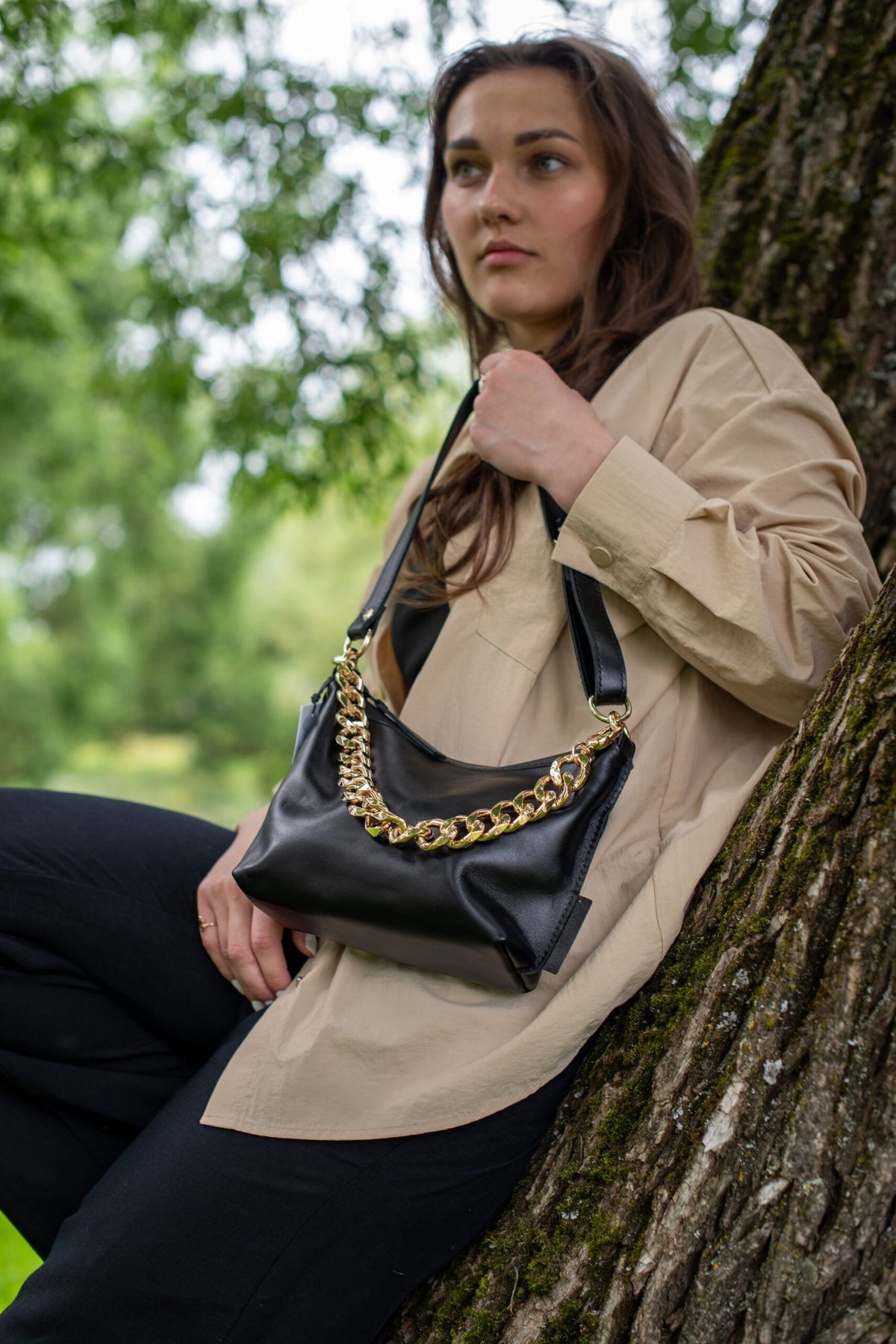 3. Woman sitting by tree with Zelma Kraft black leather hobo bag featuring gold chain