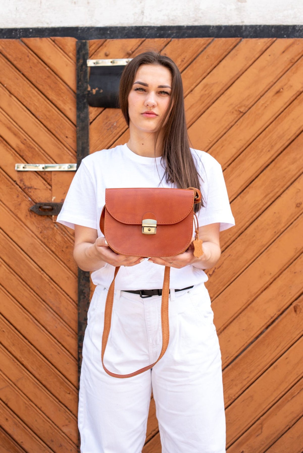1. Woman holding Zelma Kraft natural leather saddle bag in light brown, showcasing adjustable strap and secure buckle closure