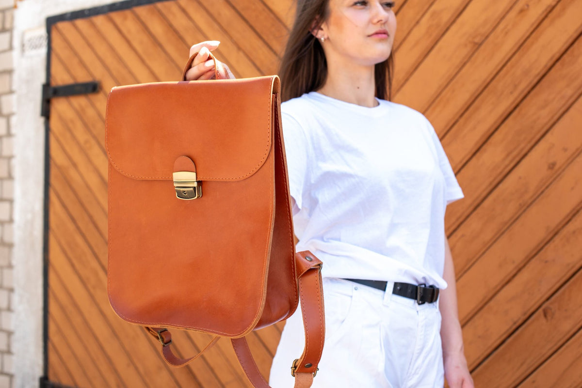 2. Woman holding Zelma Kraft light brown leather saddle backpack, showcasing front design and clasp in lifestyle setting