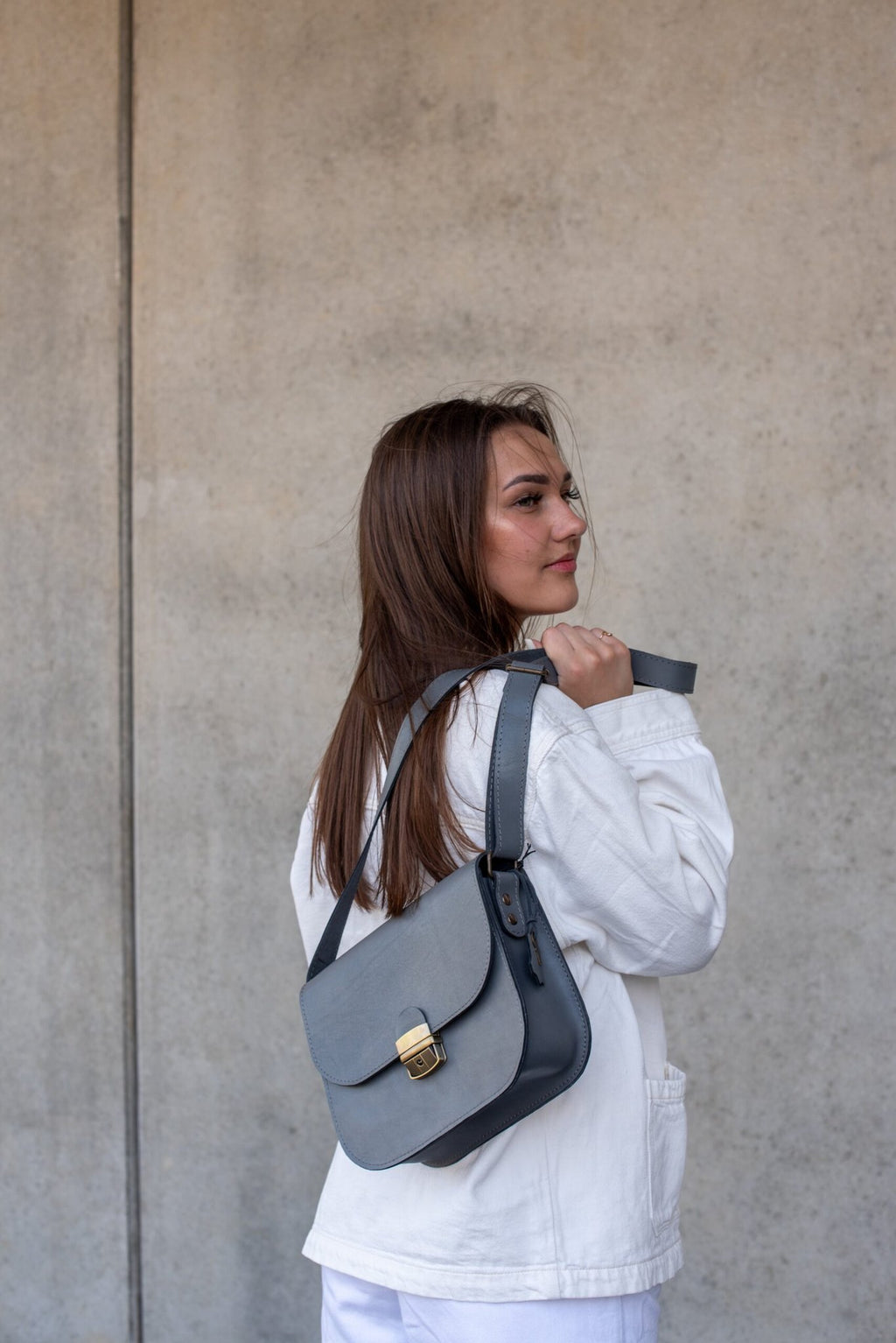 1. Woman wearing Zelma Kraft grey leather saddle bag with adjustable strap, styled with white jacket