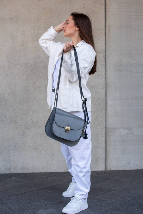 5. Woman adjusting the strap of the Zelma Kraft grey leather saddle bag, styled with a white outfit, against a concrete wall