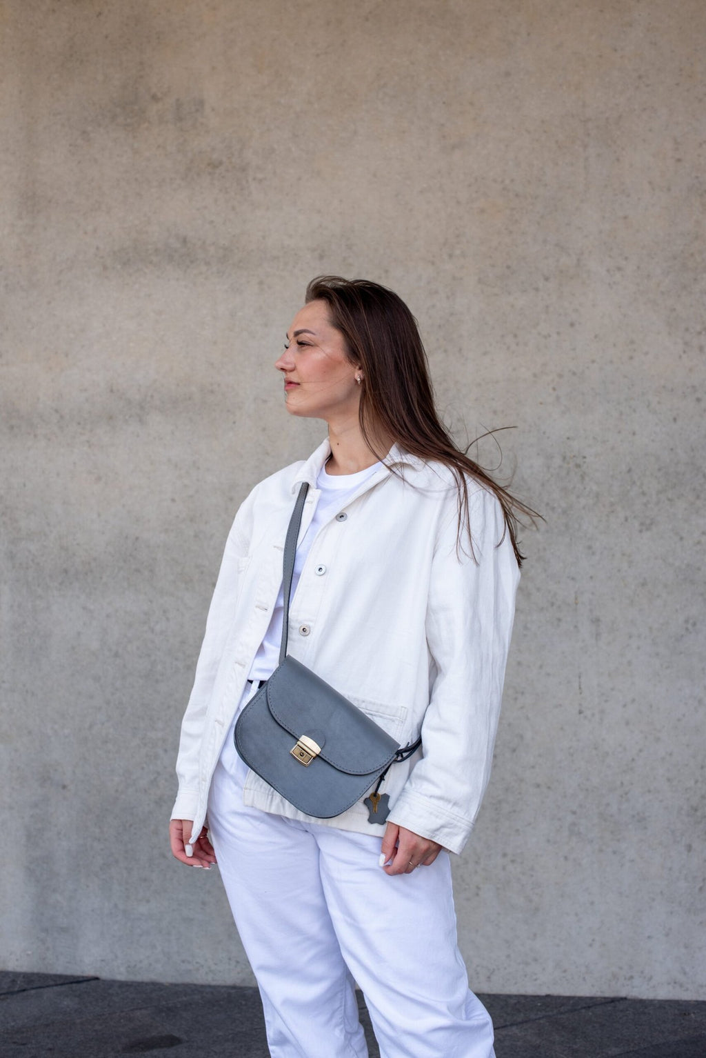 4. Woman wearing the Zelma Kraft grey leather saddle bag crossbody, styled with a white outfit, against a concrete background