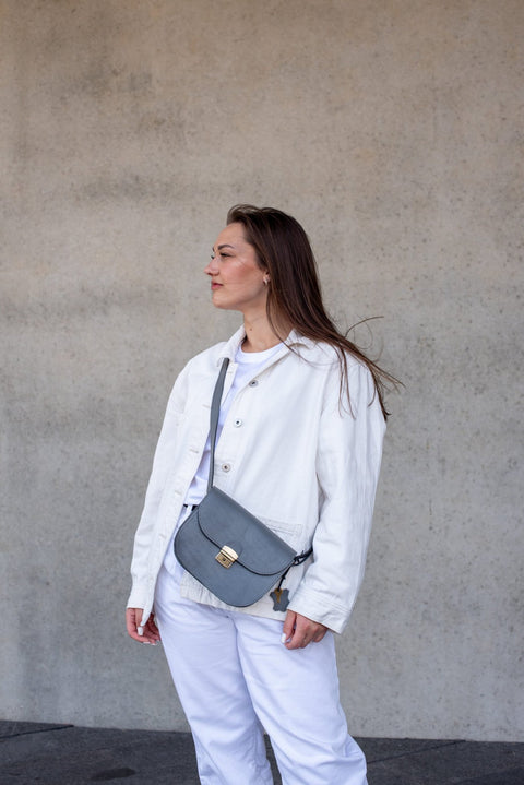 4. Woman wearing the Zelma Kraft grey leather saddle bag crossbody, styled with a white outfit, against a concrete background