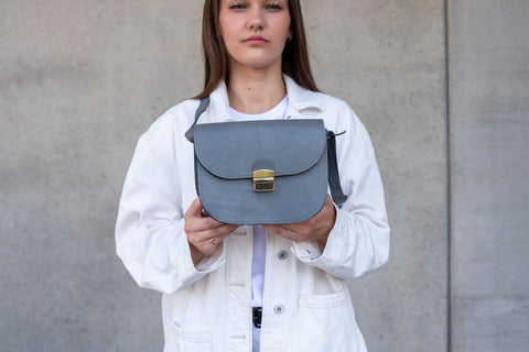 3. Woman holding the Zelma Kraft grey leather saddle bag in front of a concrete wall, showcasing its size and style