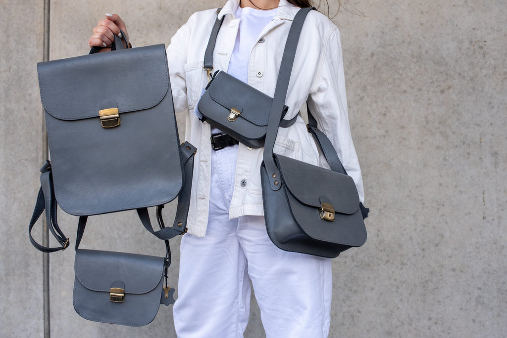 6. Woman holding multiple Zelma Kraft grey leather bags, including the saddle bag, styled with a white outfit, against a concrete wall