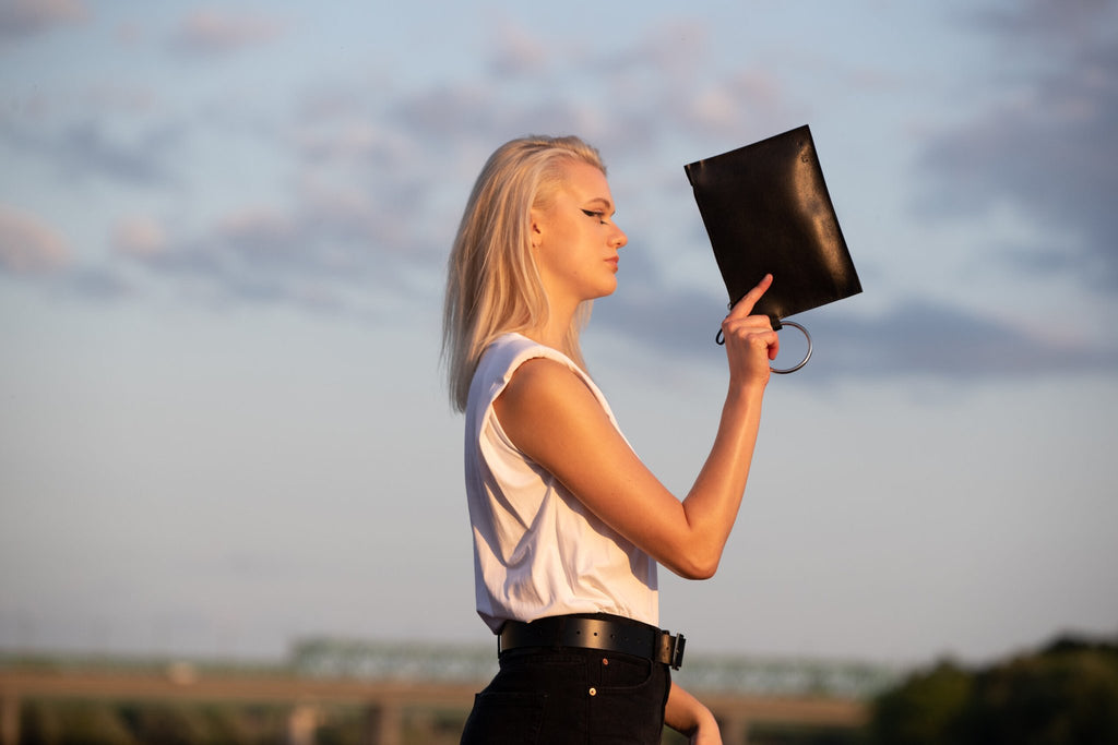 2. Woman posing with PYKOK Clutch Nina, highlighting black leather and metal ring handle against a scenic backdrop