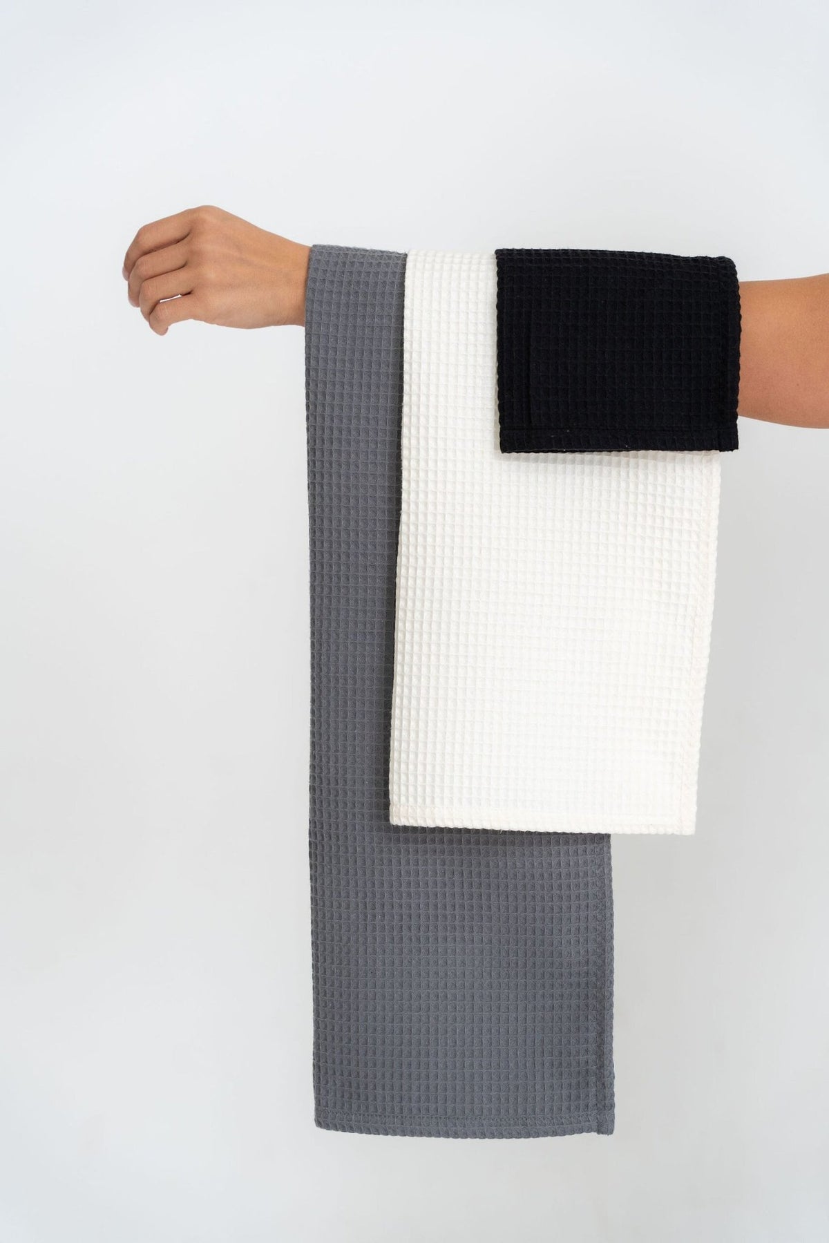 2. Arm holding three folded waffle cotton towels in gray, white, and black against a white background