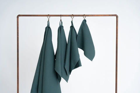 1. Set of four petrol organic waffle cotton towels hanging on a copper rack against a white background
