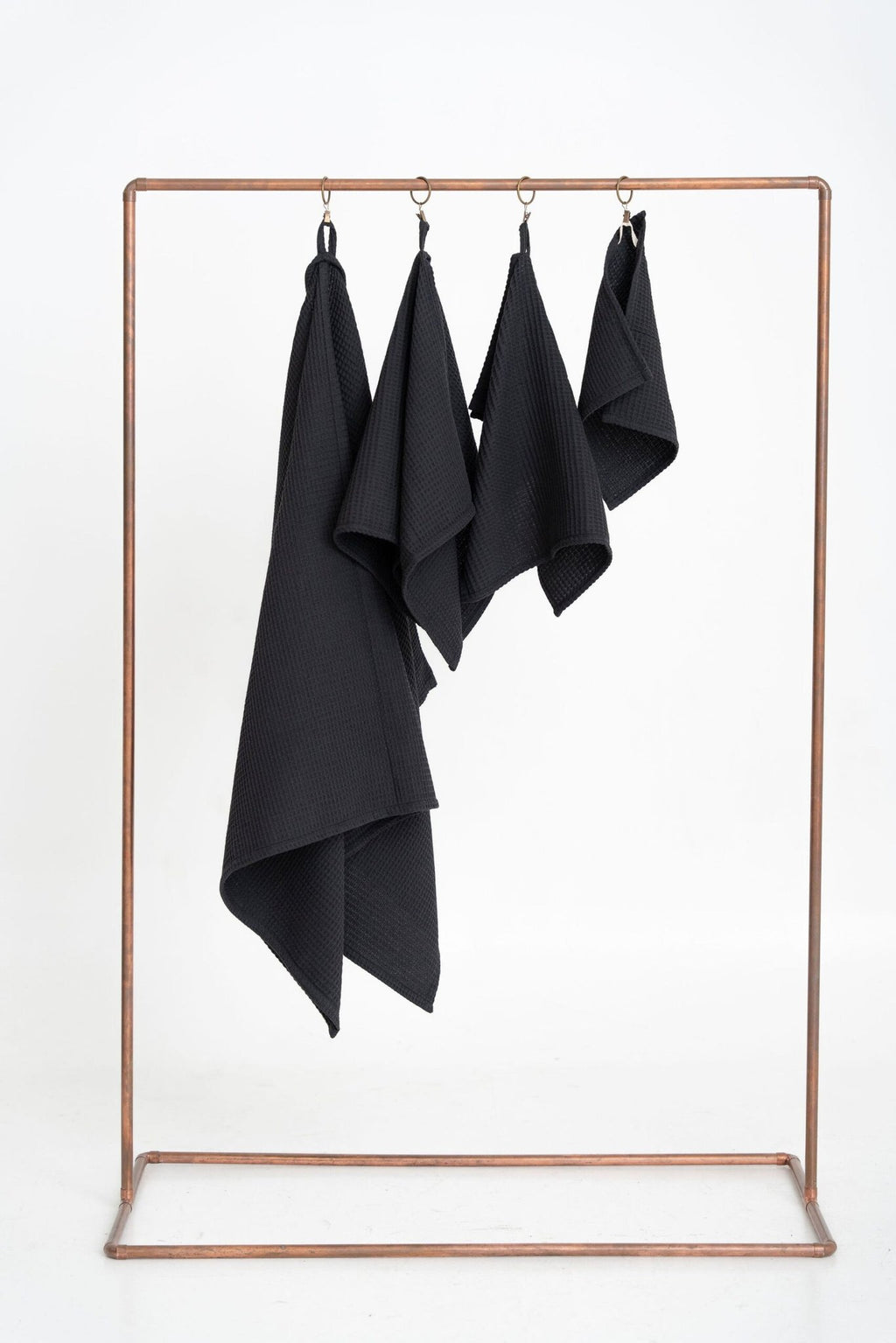 1. Set of four black organic waffle cotton towels hanging on a copper rack against a white background