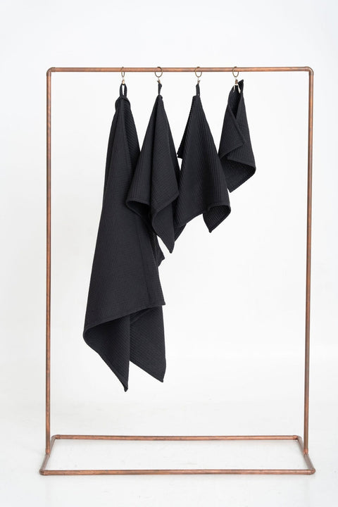 1. Set of four black organic waffle cotton towels hanging on a copper rack against a white background