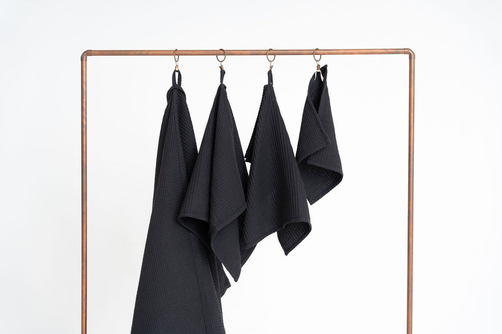 4. Four black waffle cotton towels hanging on a copper rack, displayed in various sizes against a white background