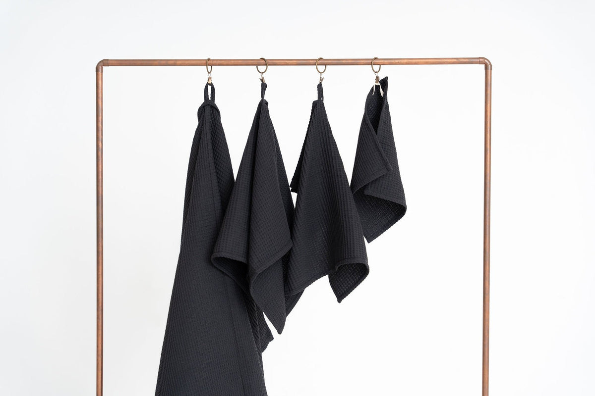 4. Four black waffle cotton towels hanging on a copper rack, displayed in various sizes against a white background