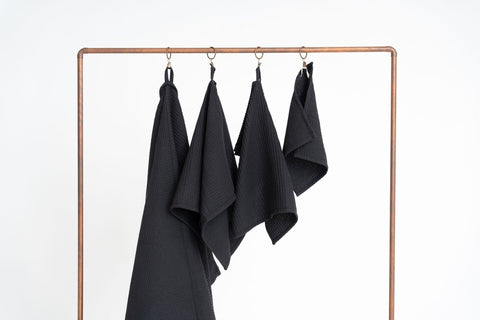 4. Four black waffle cotton towels hanging on a copper rack, displayed in various sizes against a white background