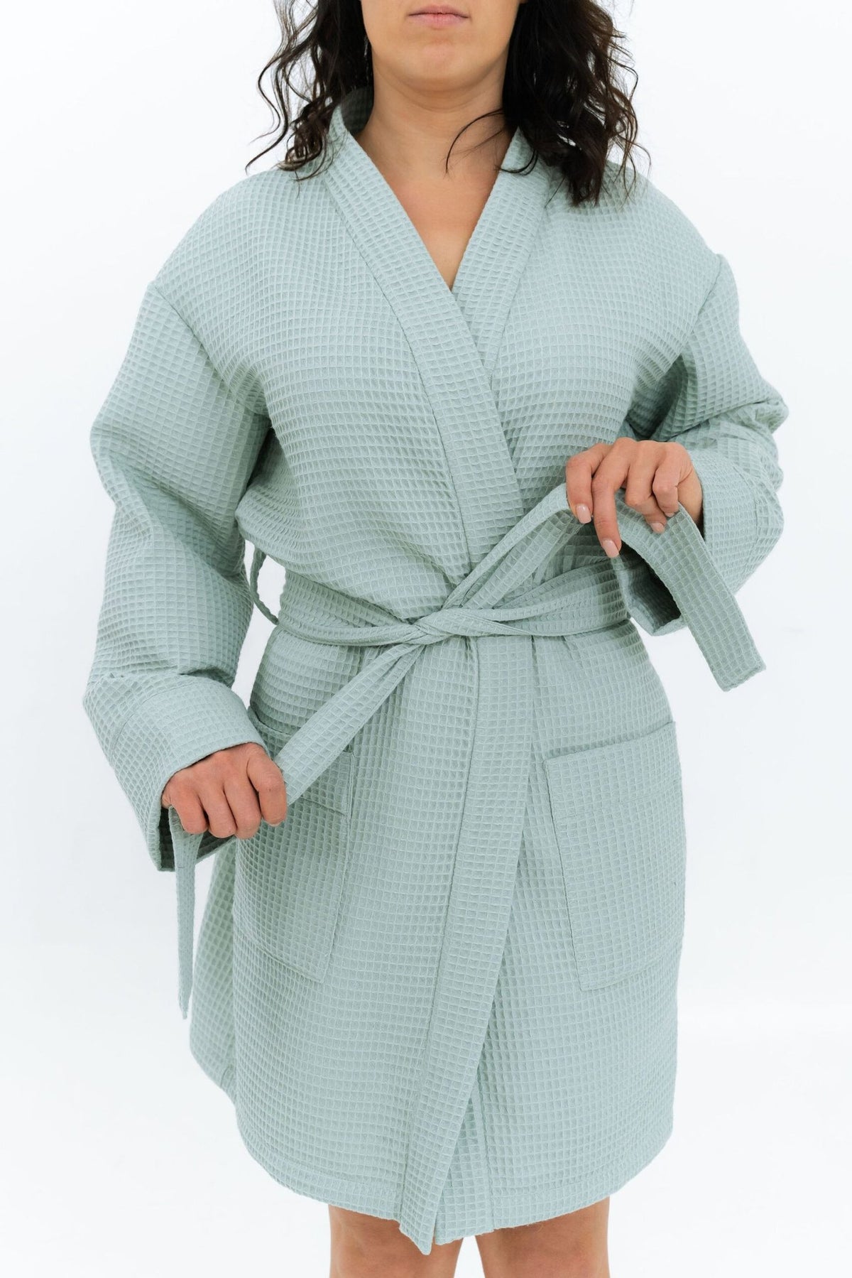 4. Woman adjusting belt of light blue waffle robe with side pockets, studio setting