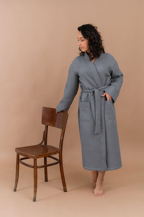 2. Woman in grey waffle robe with belt, standing next to a wooden chair, on a beige background