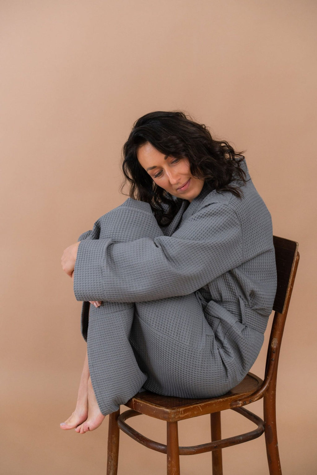 1. Woman sitting on chair wearing grey waffle fabric robe with belt, in a relaxed pose against a beige background