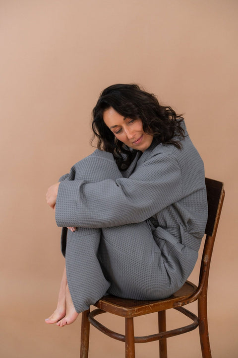 1. Woman sitting on chair wearing grey waffle fabric robe with belt, in a relaxed pose against a beige background