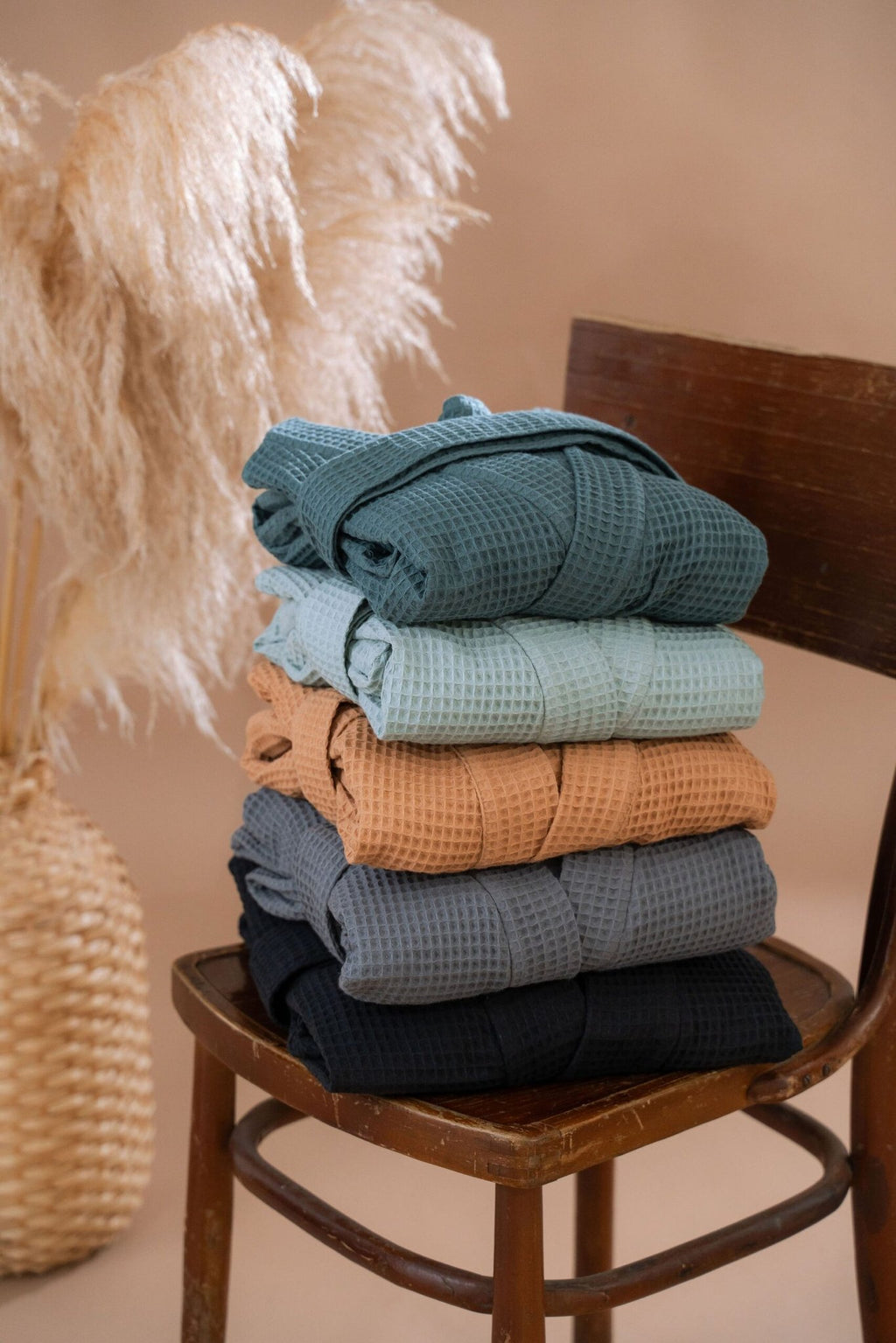 4. Stack of folded waffle robes in various colors on wooden chair with beige background