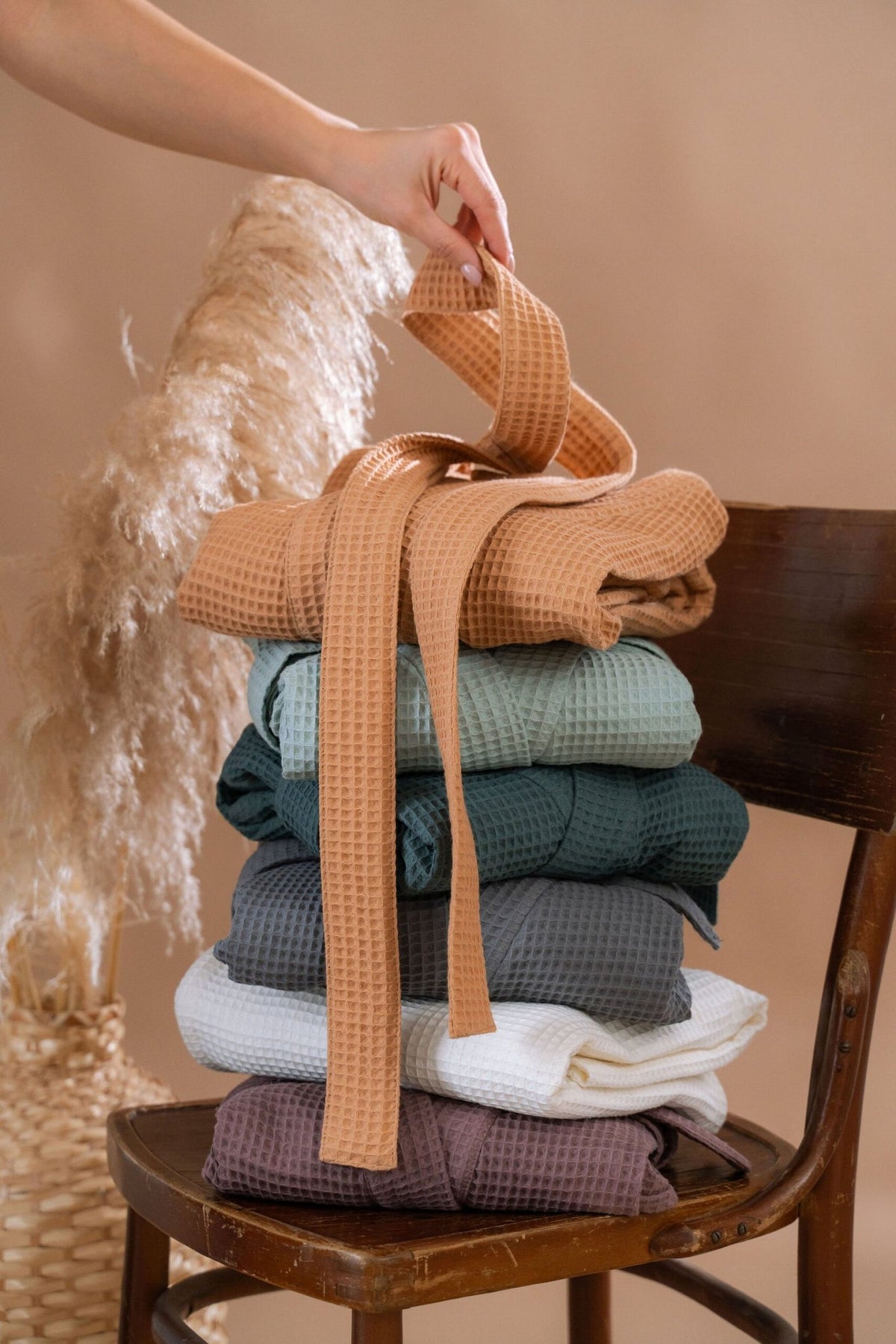 4. Stack of folded waffle fabric robes in various colors on a wooden chair, with a hand holding a belt