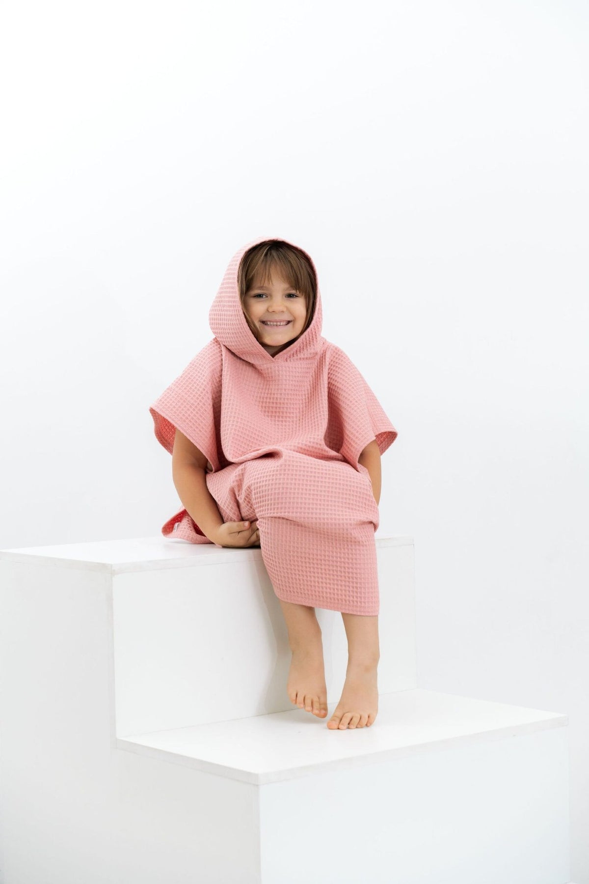 1. Child wearing pink waffle fabric poncho with hood sitting on white steps