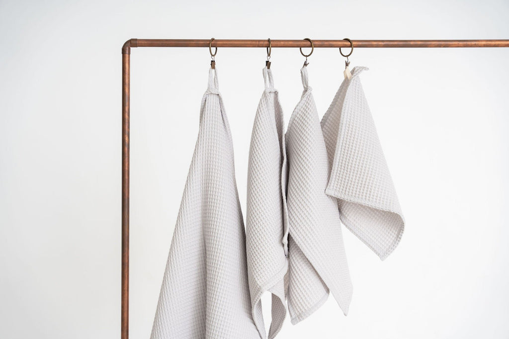 1. Set of four light gray organic waffle cotton towels hanging on copper rod, showcasing different sizes with hanging loops