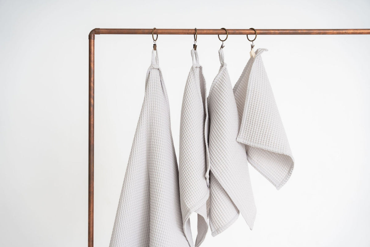 1. Set of four light gray organic waffle cotton towels hanging on copper rod, showcasing different sizes with hanging loops