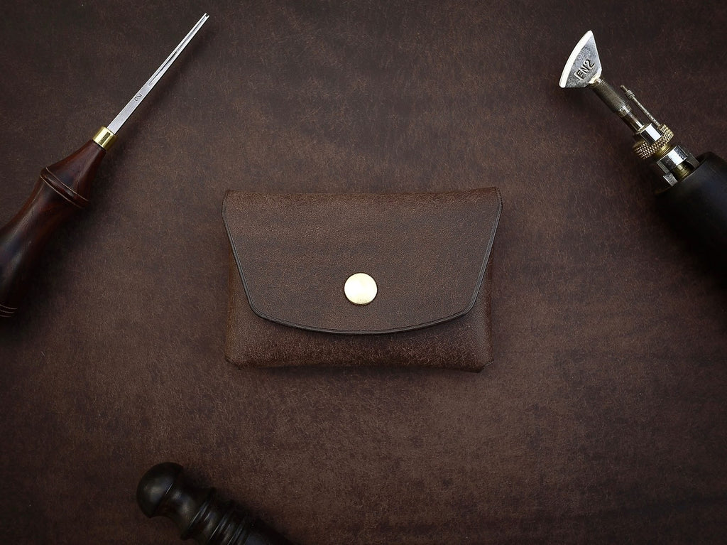 1. Mirgoods dark brown button leather wallet with brass snap, shown in a flat lay with leather tools