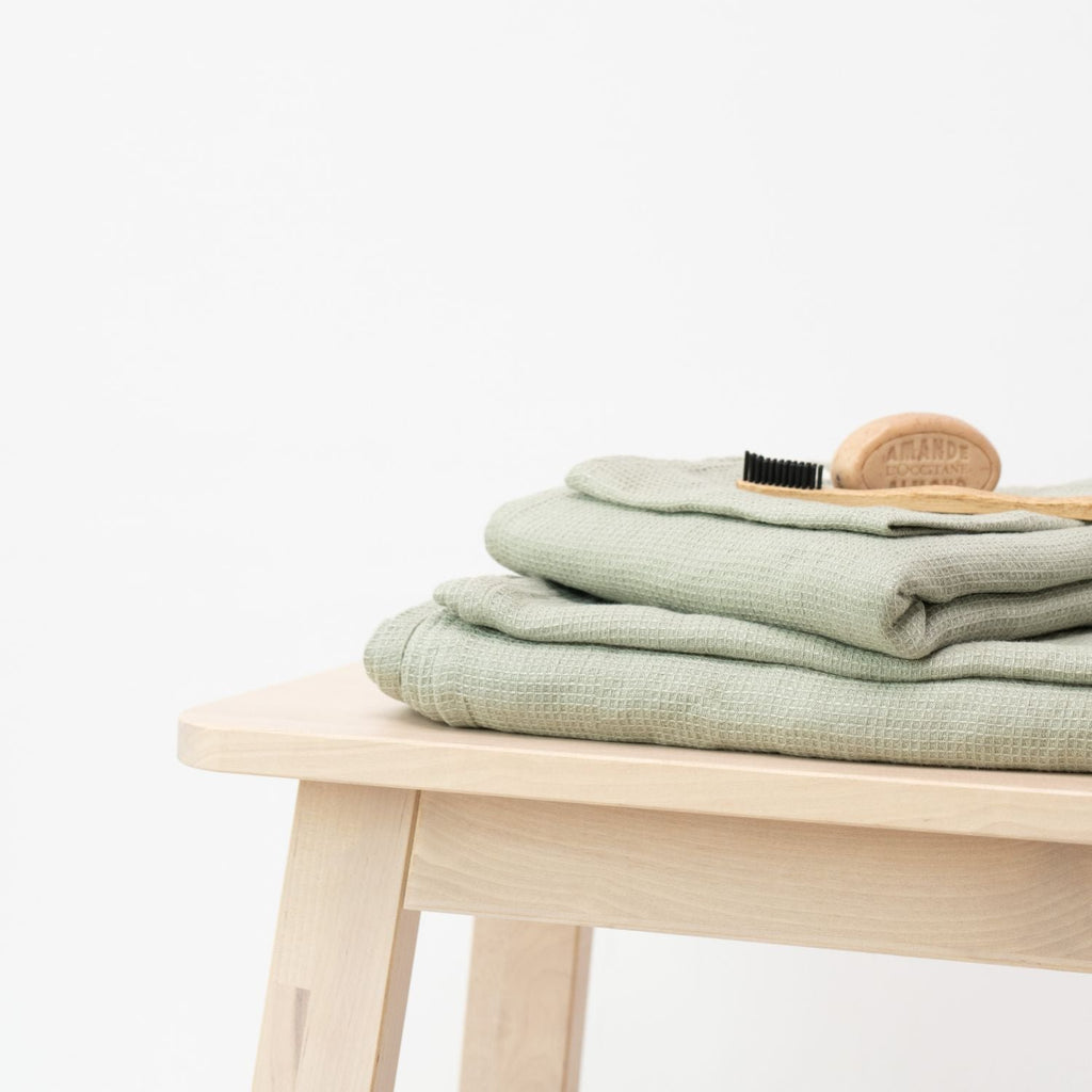 1. Sage green waffle linen towels stacked on a light wooden stool with a grooming brush on top