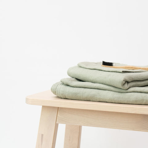 2. Sage green waffle linen towels neatly folded on a wooden stool with a grooming brush
